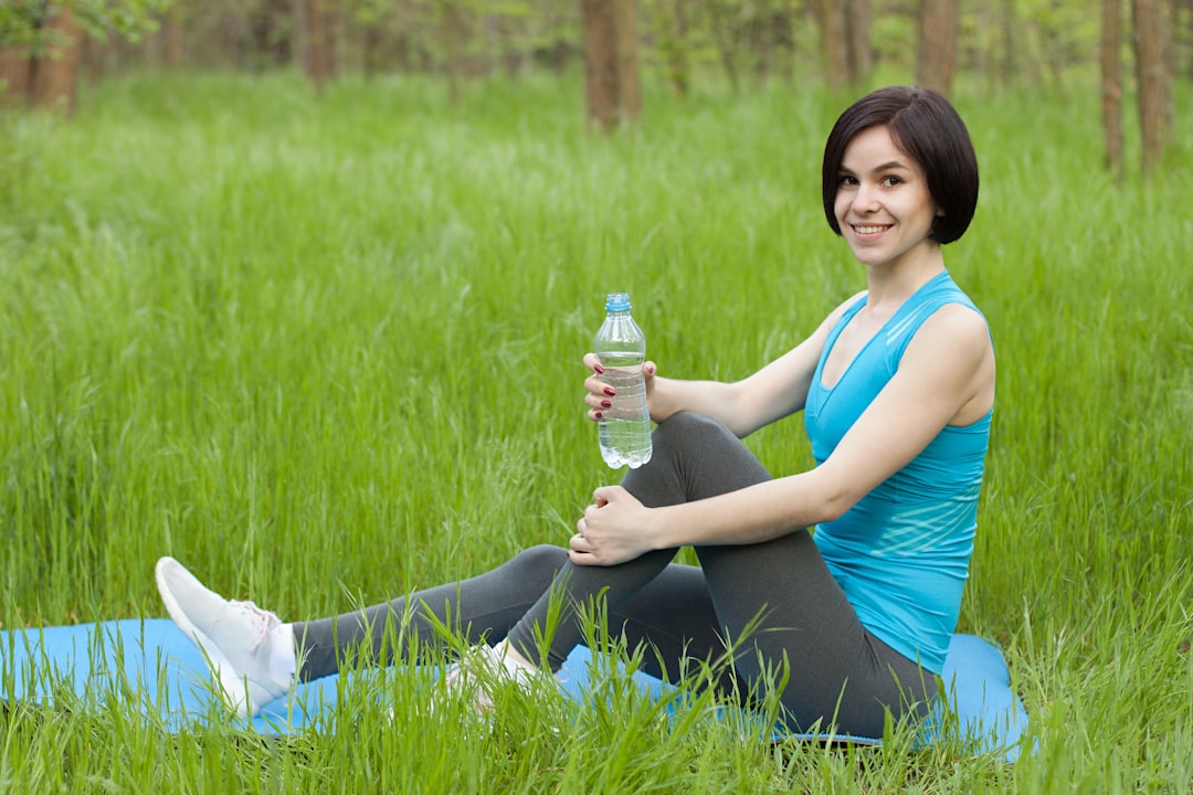 A smiling woman in fitness attire sitting on a blue mat in a grassy field, holding a bottle of water.