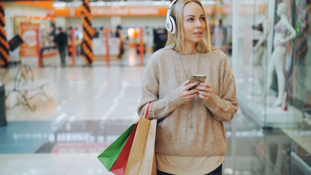 A young blonde woman wearing a beige sweater and white over-ear headphones stands in a brightly lit shopping mall, holding a smartphone and several paper shopping bags (red, green, and brown). S
